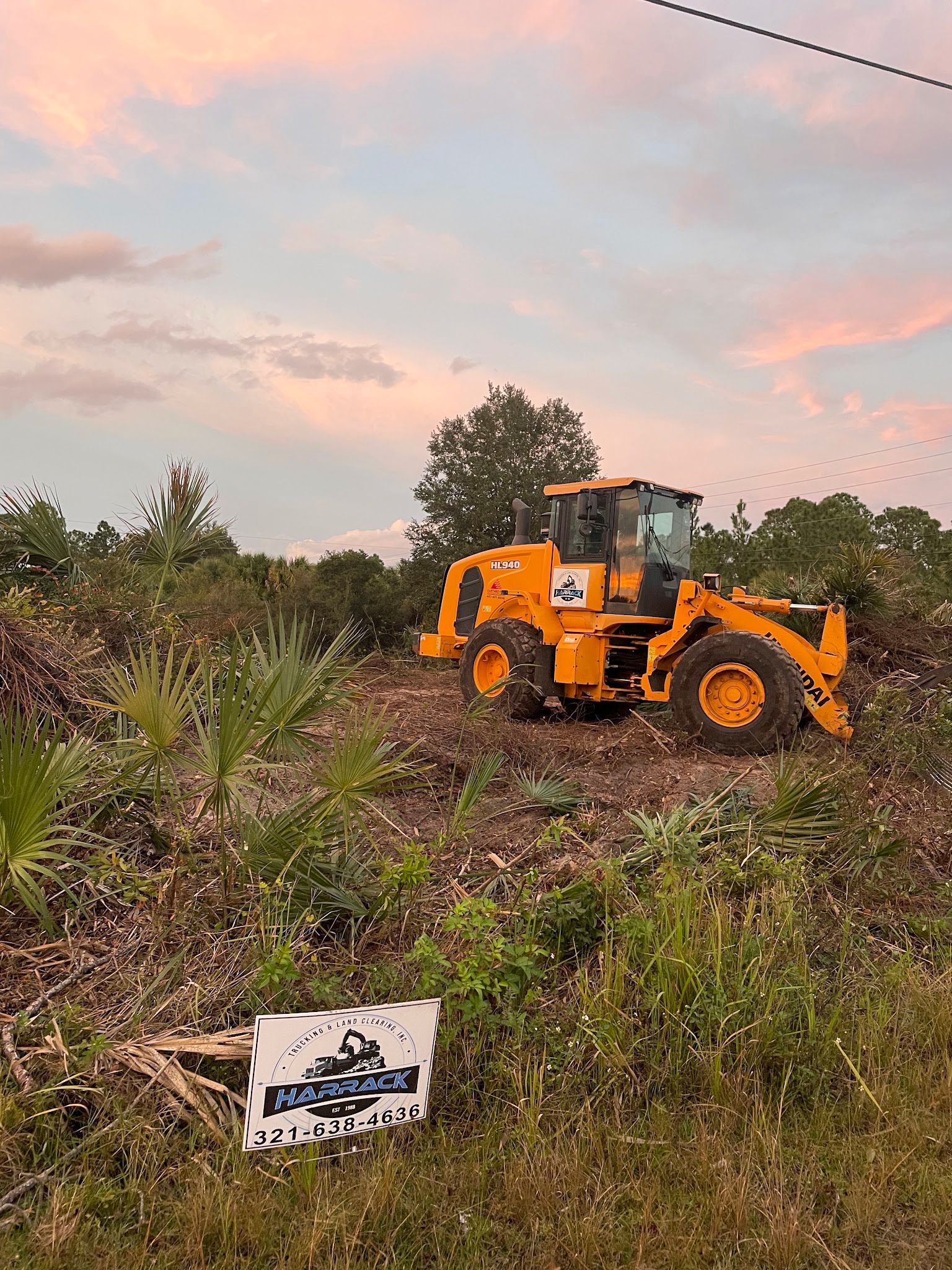 Yellow wheel loader clearing palmetto at sunset, Harrack sign in front